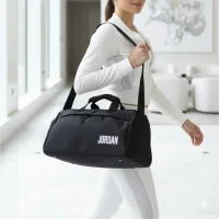 کیف ورزشی مشکی با نوشته Jordan روی دوش یک خانم با استایل ورزشی در یک فضای داخلی لوکس و روشن، راهرویی سفید با پنجره‌های بزرگ و مبلمان مدرن.